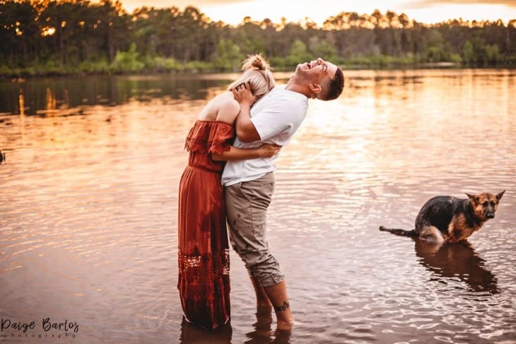 a smiling man and woman stand in the water while a dog poops in the background