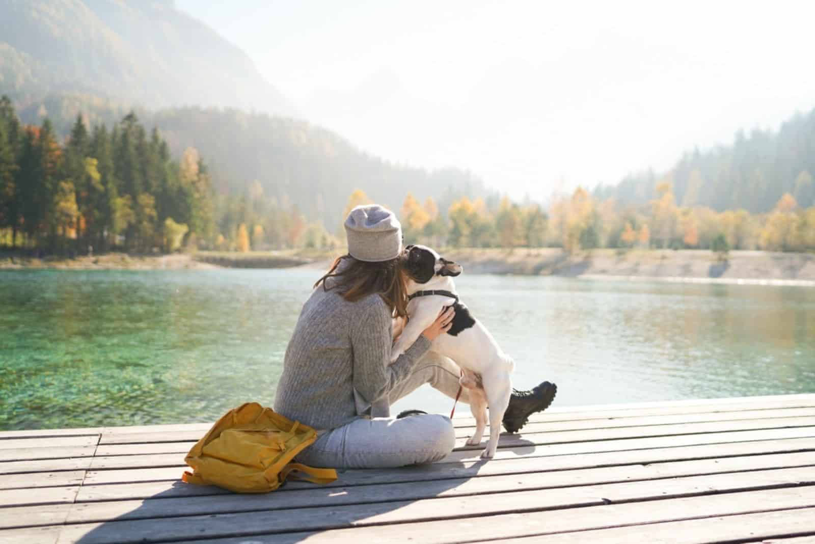 a woman sits on a pier while a French bulldog jumps on her