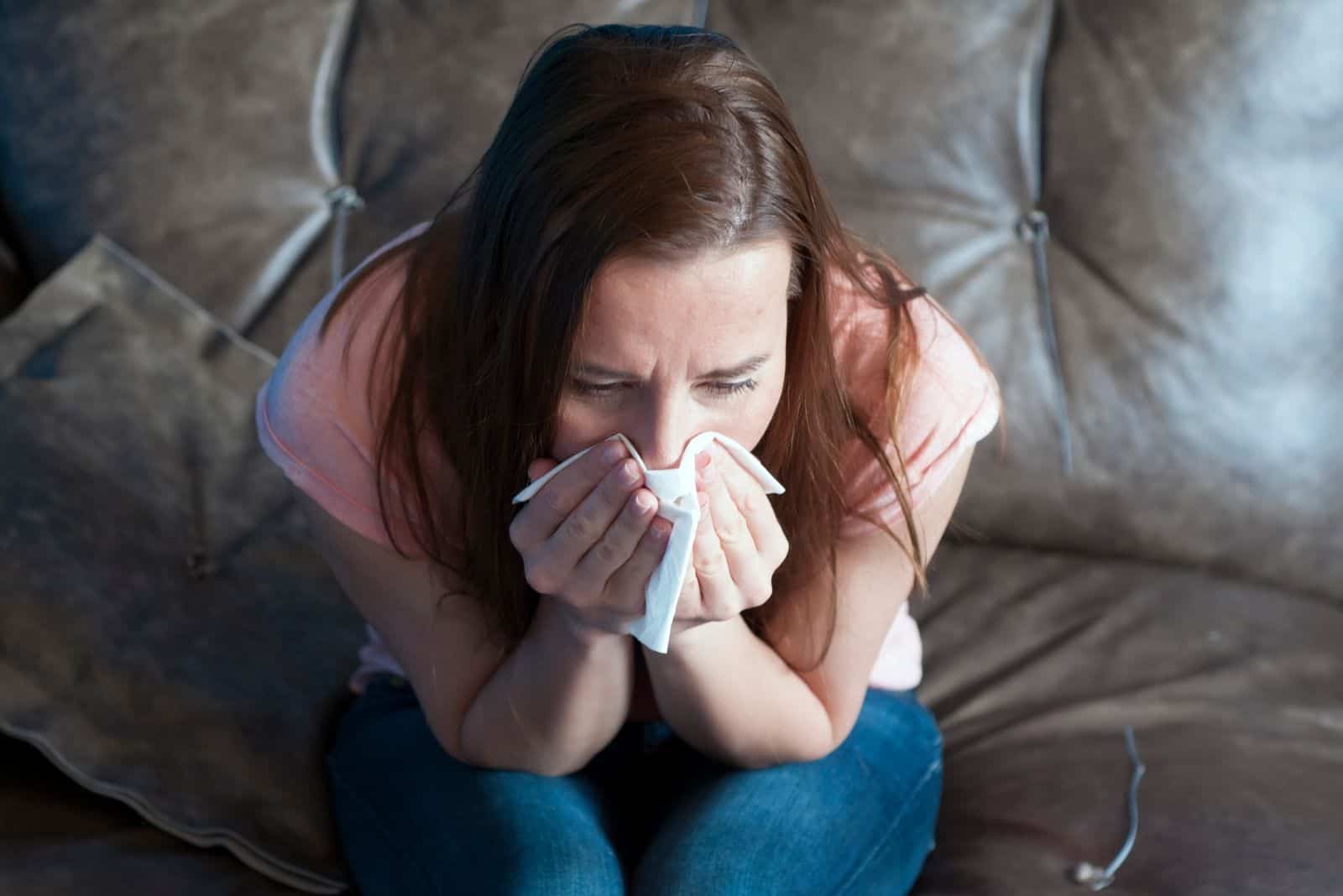 young woman sneezing on the hanky while sitting on the sofa on top angle