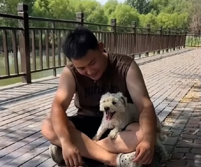 a smiling man sits on a wooden platform and a dog sits on his lap